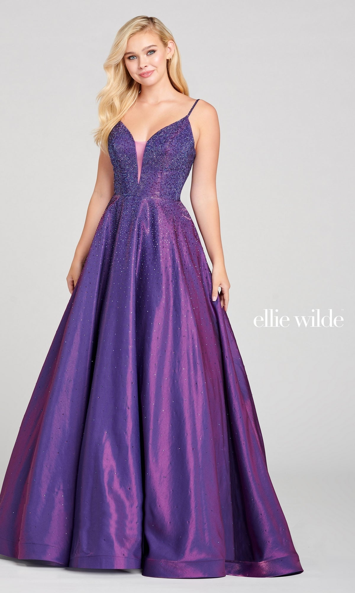 A woman in the Ellie Wilde Ew121005 Long Beaded Prom Ball Gown, a shimmering purple A-line dress with thin straps and a full skirt, stands against a plain gray background. The text